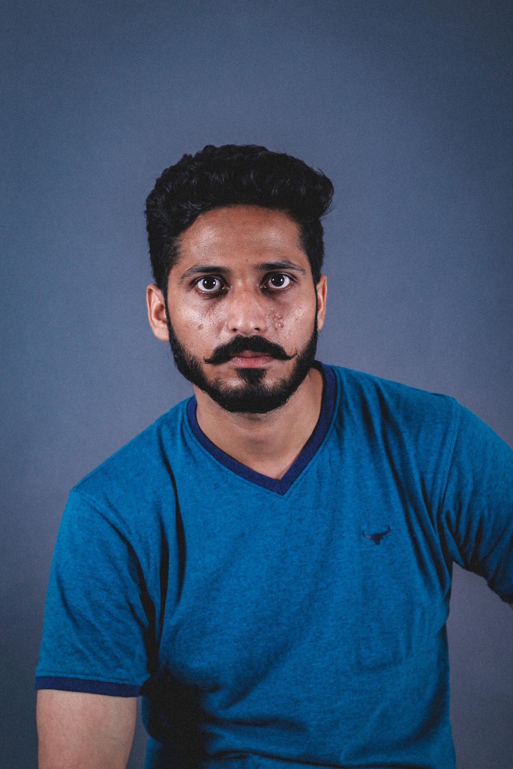 A man with facial hair in a blue shirt poses for a portrait against a gray background.